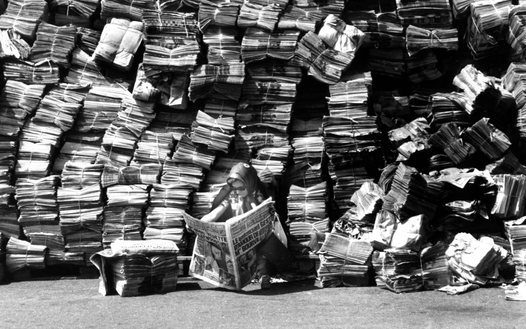Woman reading a newspaper in front of a big pile of newspapers.