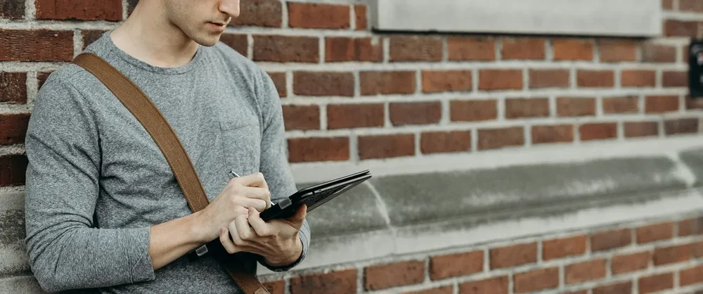 En man i grå tröja står lutad mot en tegelvägg och skriver på sin surfplatta.