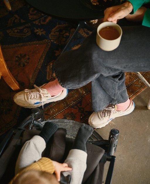 En vuxen person sitter med korslagda ben och dricker te ur en kopp. Mittemot sitter ett barn i en barnvagn.