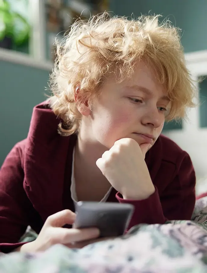 Pojke ligger i sängen med mobilen och ser fundersam ut. Kampen mot näthat.
