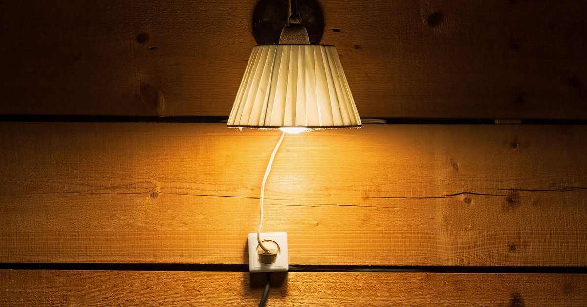 A lit lamp hanging on a wooden wall, plugged in slightly underneath where it’s hanging. The lampshade is white and crinkly, with a farmhouse feel to it.