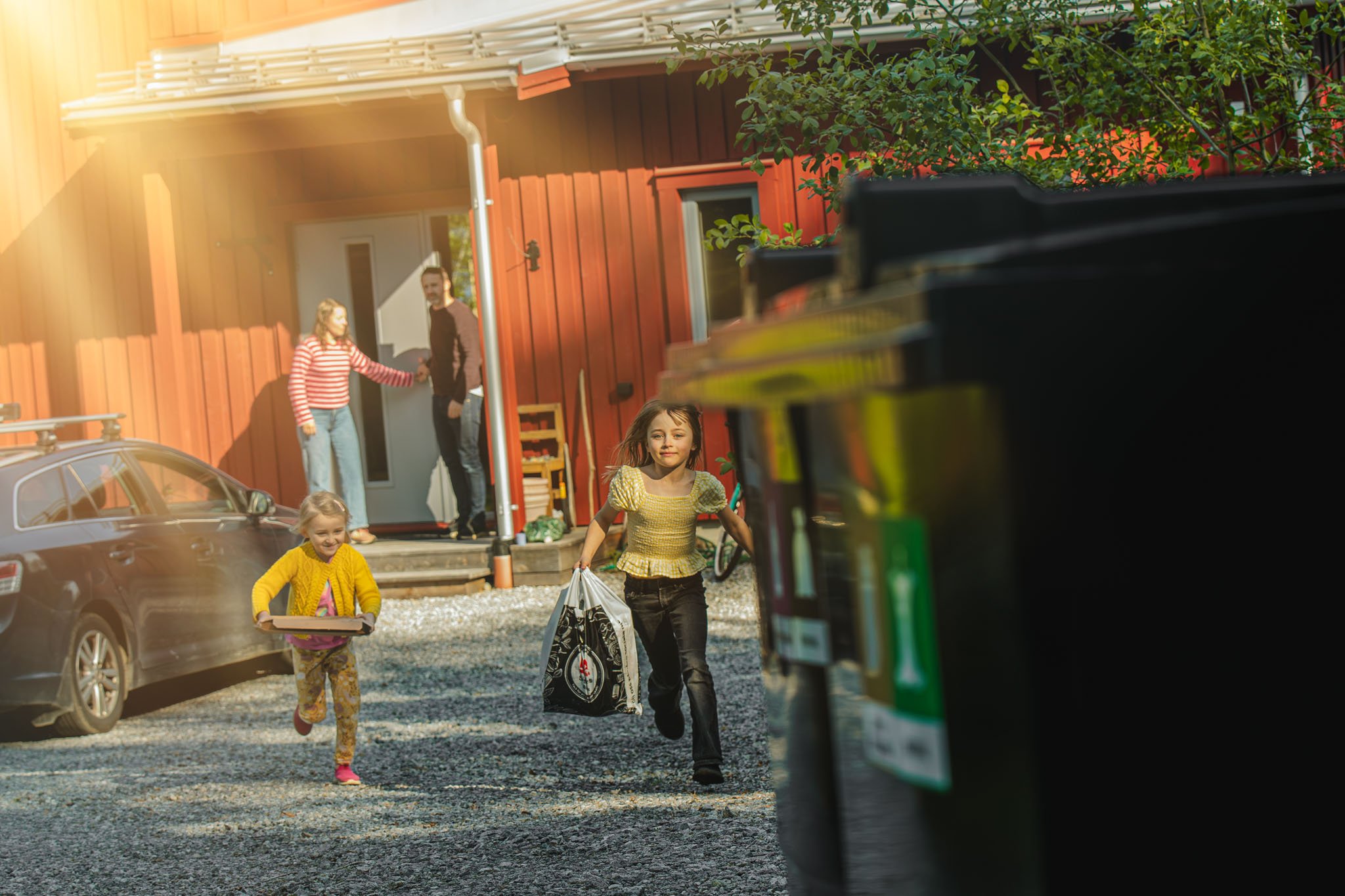 Tv&aring; barn springer i en g&aring;rd framf&ouml;r ett r&ouml;tt hus. En vuxen st&aring;r i d&ouml;rren. Bil och gr&ouml;nska syns.