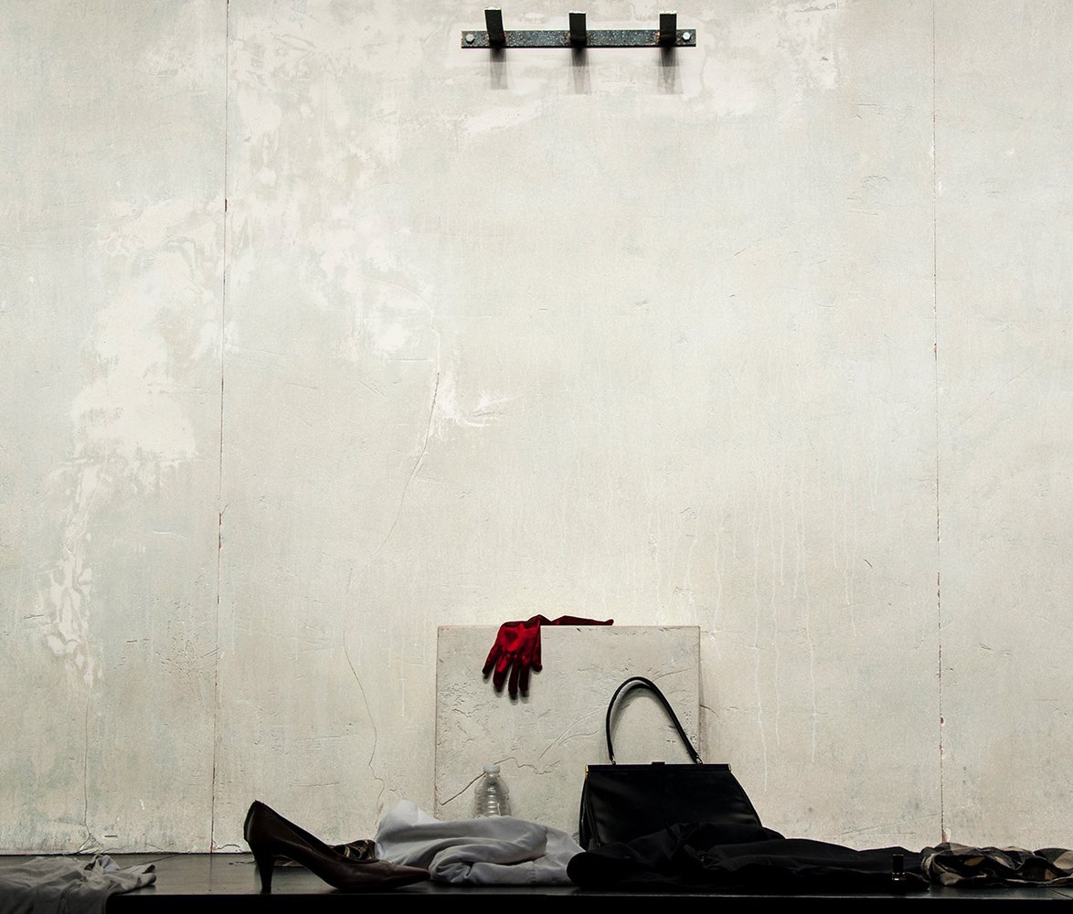 En vit vägg med tre krokar, nedanför på golvet ligger en väska, klackskor och några kläder.