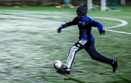 Pojke i mössa och vantar springer med en fotboll i i svagt elljussken.