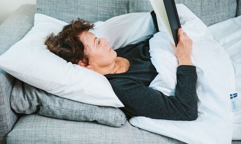 Kvinna som ligger i soffa med tyngdtäcke över sig och läser en bok