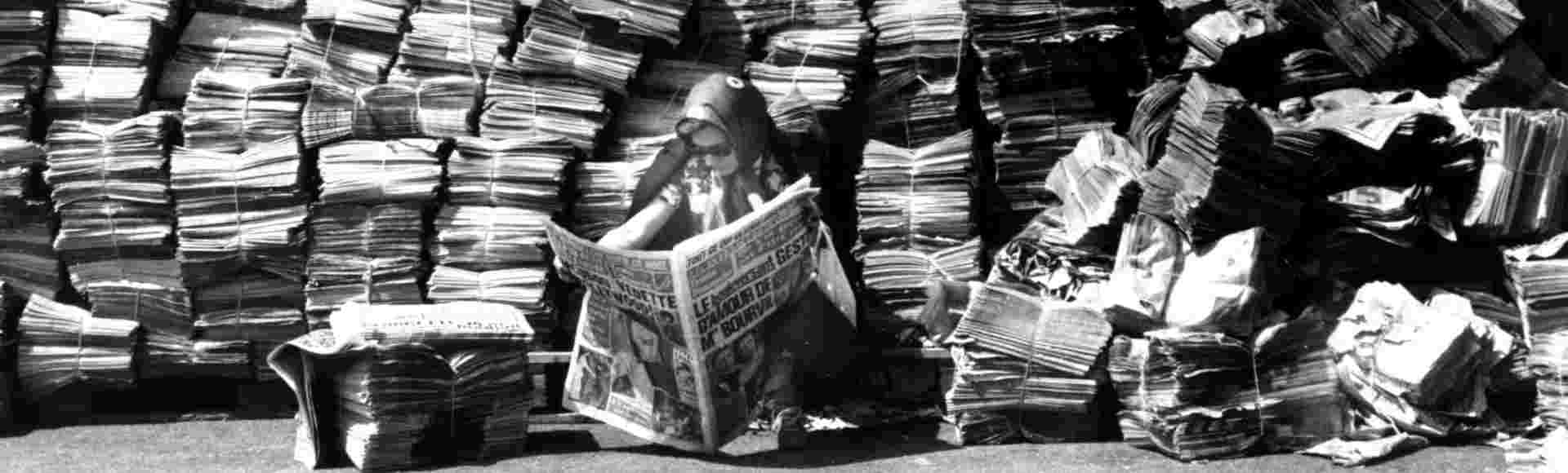 Woman reading a newspaper in front of a big pile of newspapers.