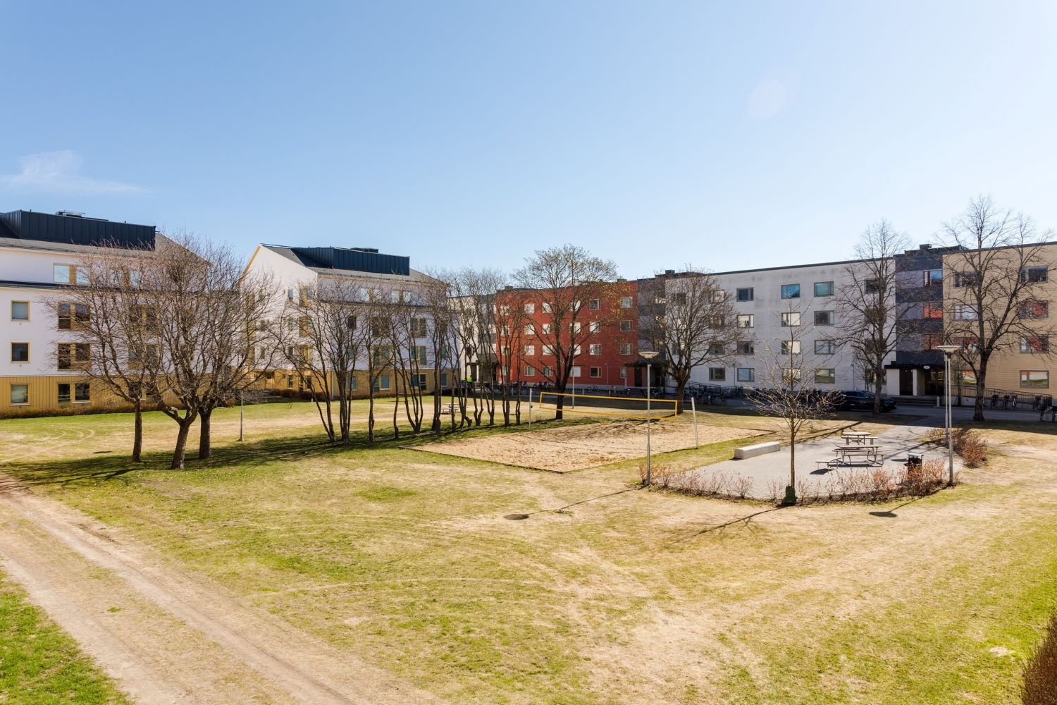 Exterior of white, yellow and red buildings, a large grass lawn and trees, a sand volleyboll court and sitting area with tables and benches.