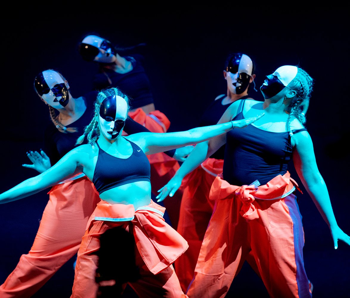 Dansgrupp uppträder på scen i blått ljus, iklädda svart scenklädsel och orange plagg runt midjan, med svartvita masker som täcker ansiktet och armarna utsträckta i synkron rörelse.