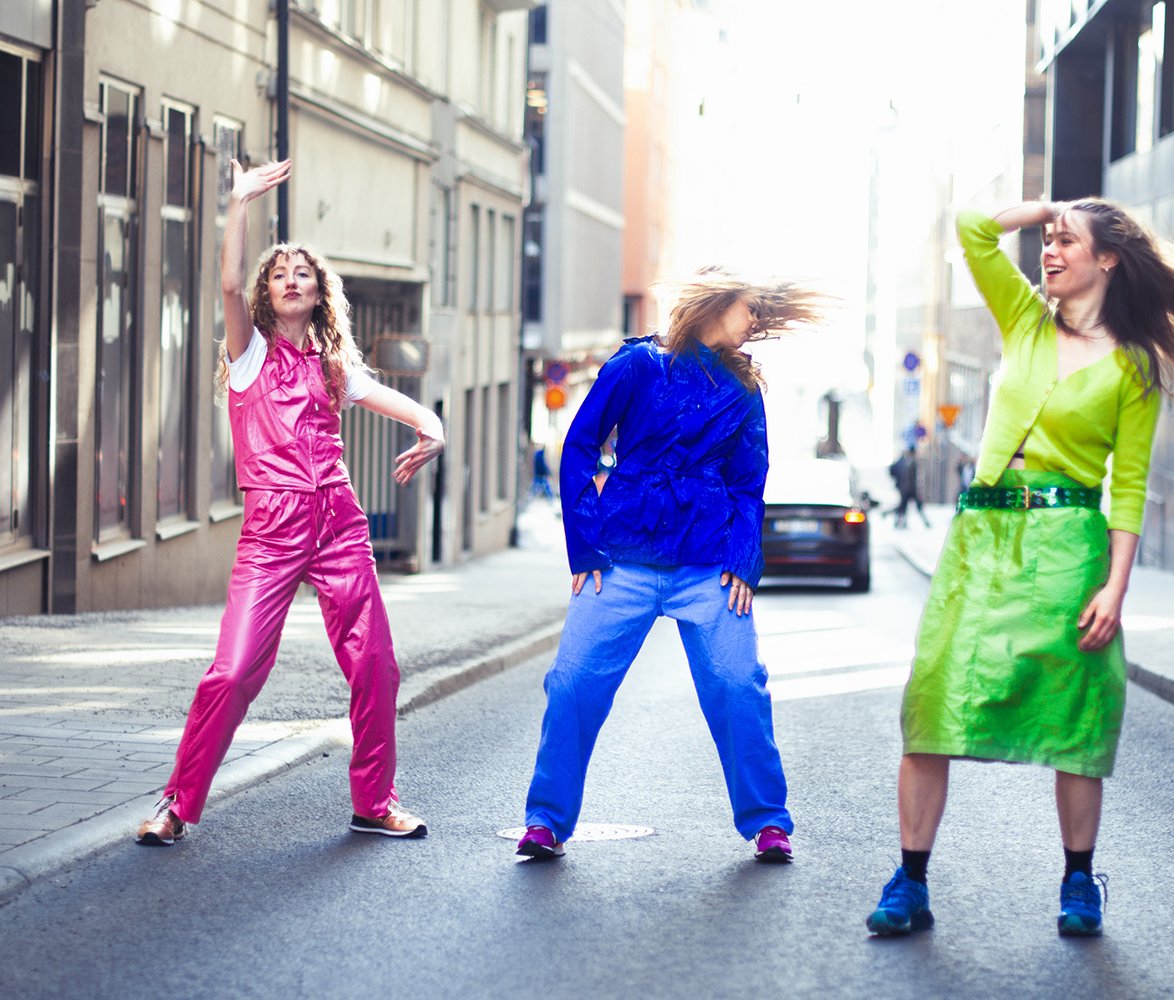 Tre kvinnor dansar mitt på en stadsgata i färgstarka kläder, omgivna av byggnader i dagsljus.
