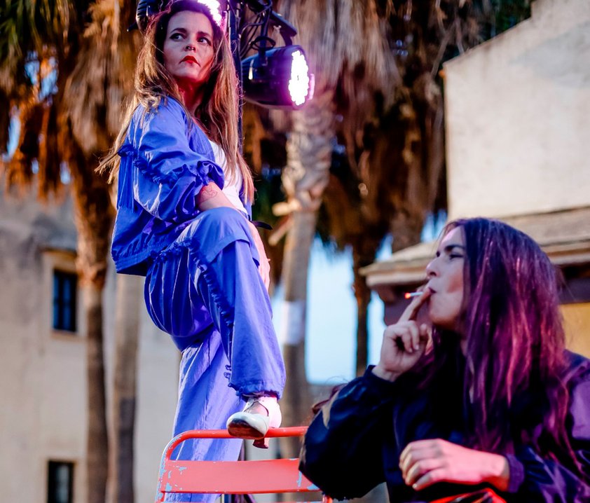 Två personer på en gård med palmer, den ena har foten på en stol, den andra röker.