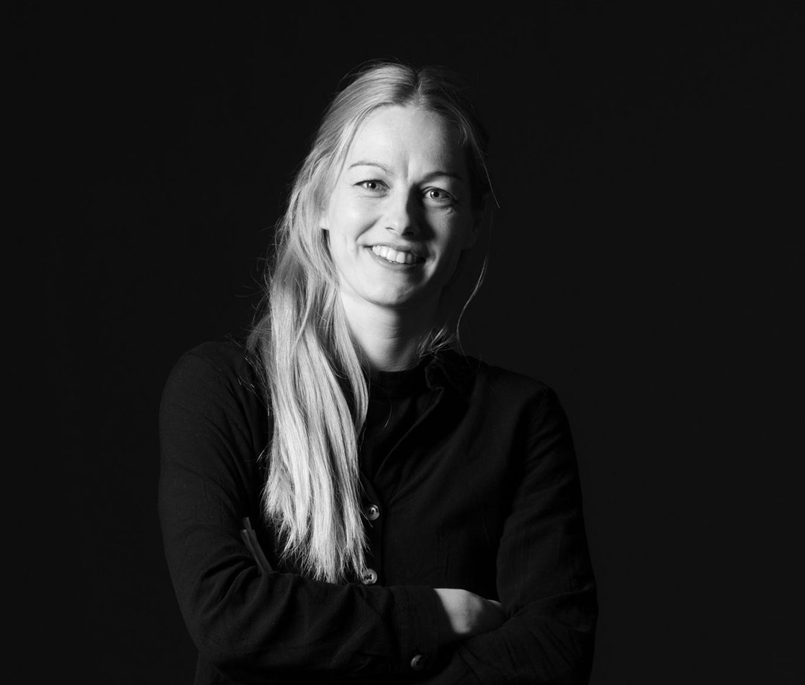 Svartvit porträttbild av en leende kvinna med långt ljust hår och armarna i kors mot en mörk bakgrund.
