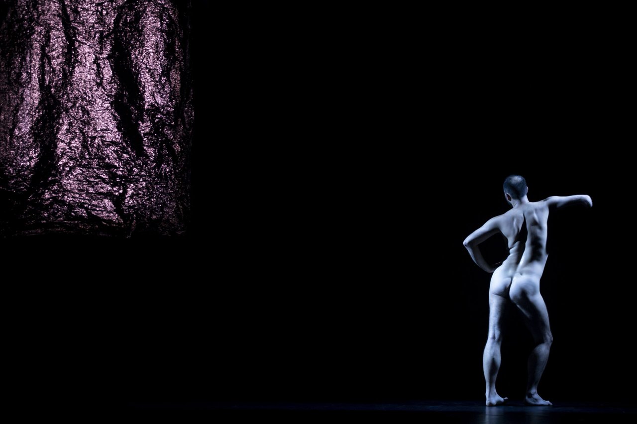 Naken dansare sedd bakifrån i en skulptural pose, upplyst mot en mörk scen med ett ljusfält i bakgrunden.