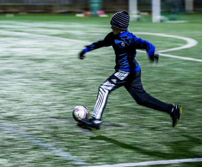 Pojke i mössa och vantar springer med en fotboll i i svagt elljussken.