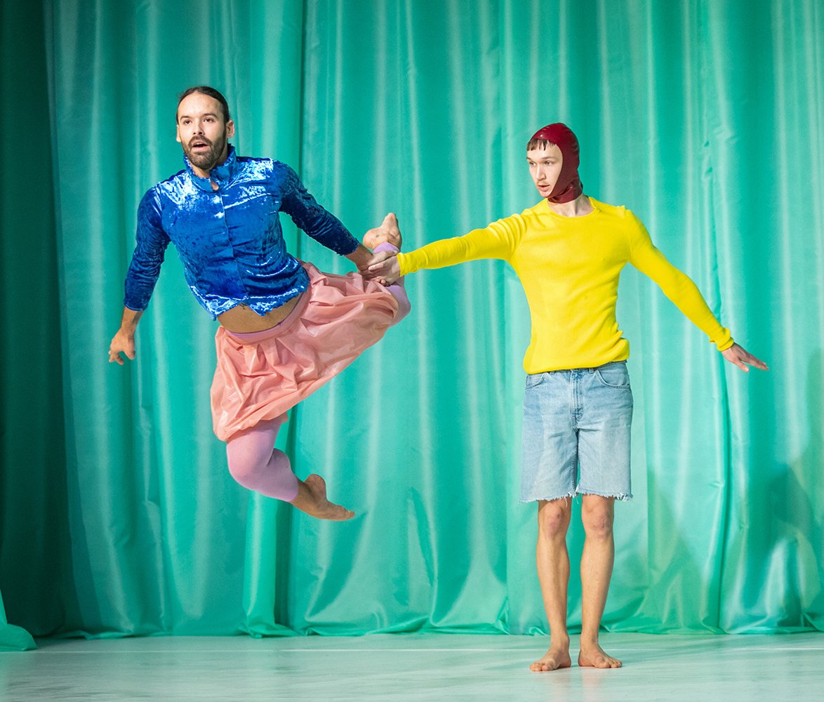 Två dansare i färgstarka kläder poserar mot en turkos bakgrund, där den ena svävar i ett hopp och den andra står stadigt.