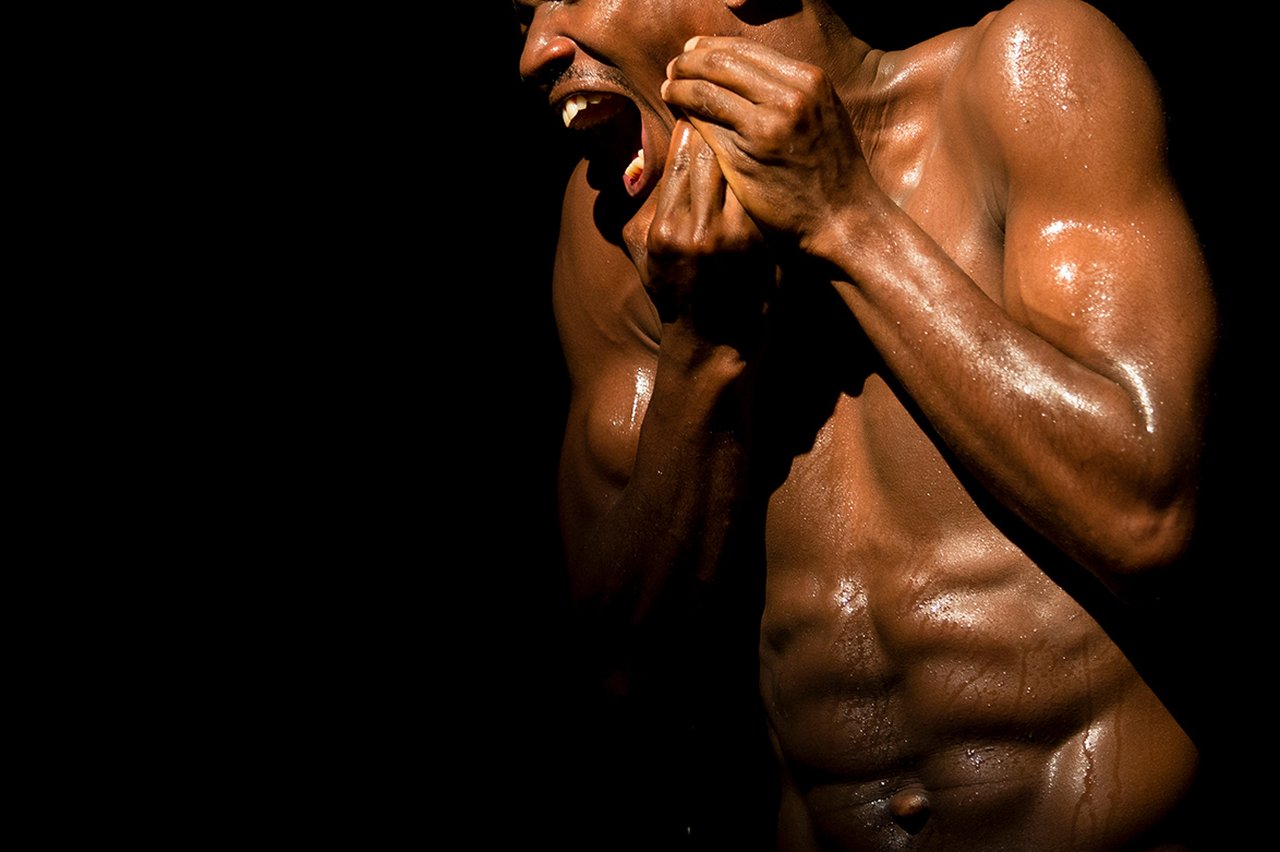 En svettig, barbröstad mörkhyad man med muskulös kropp uttrycker stark känsla genom ansikte och händer mot helt svart bakgrund.