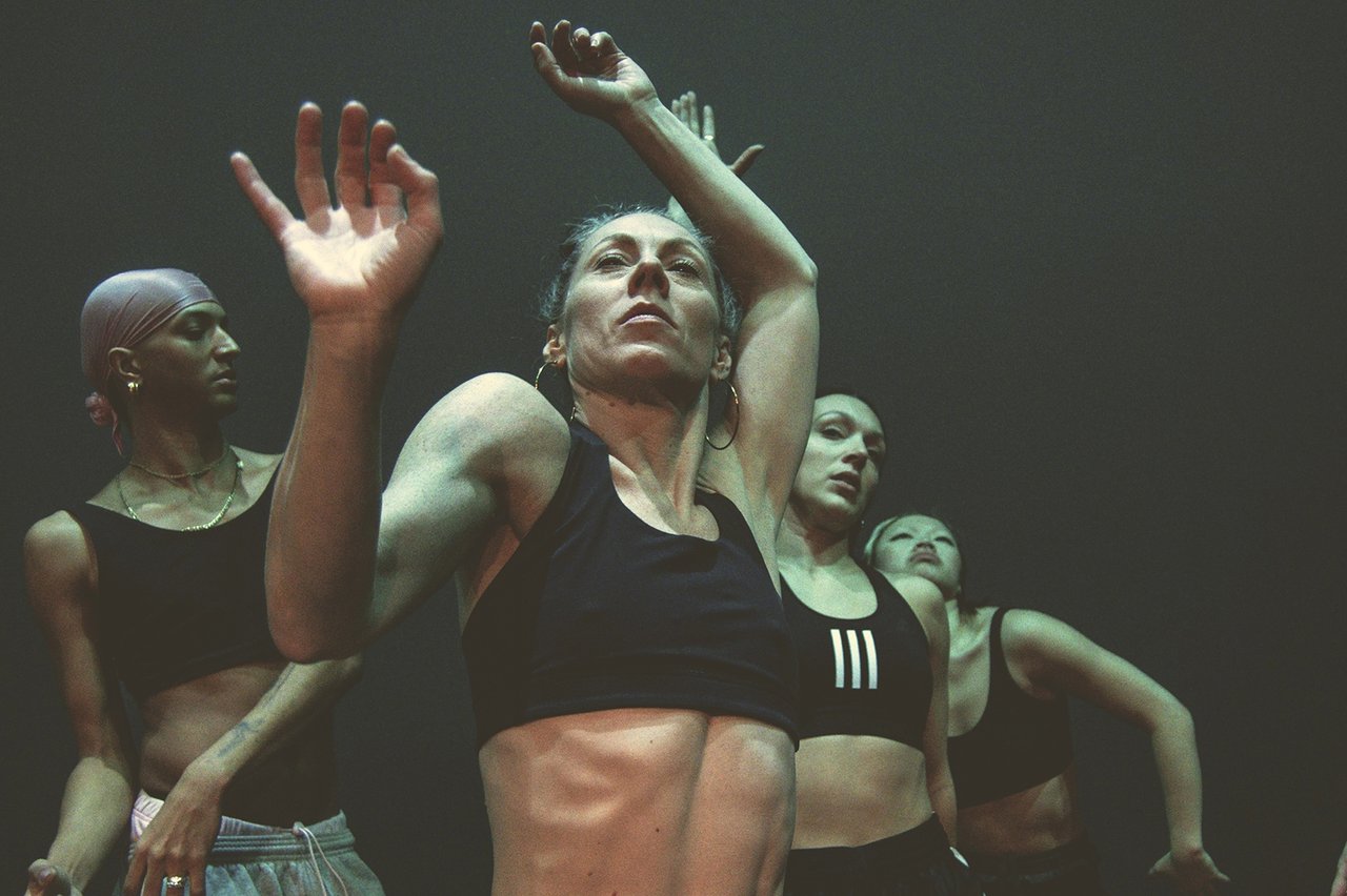 Grupp av dansare i synkron rörelse, med händer lyfta och fokus framåt i dämpat ljus