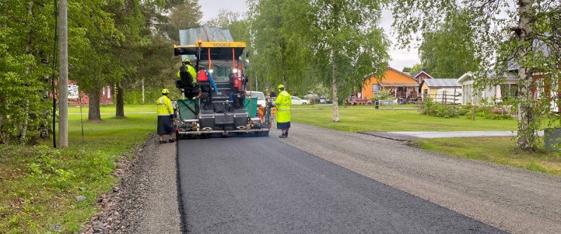 V&auml;garbet med asfaltl&auml;ggare och arbetare i gula v&auml;star p&aring; landsv&auml;gv&auml;g omgiven av tr&auml;d.