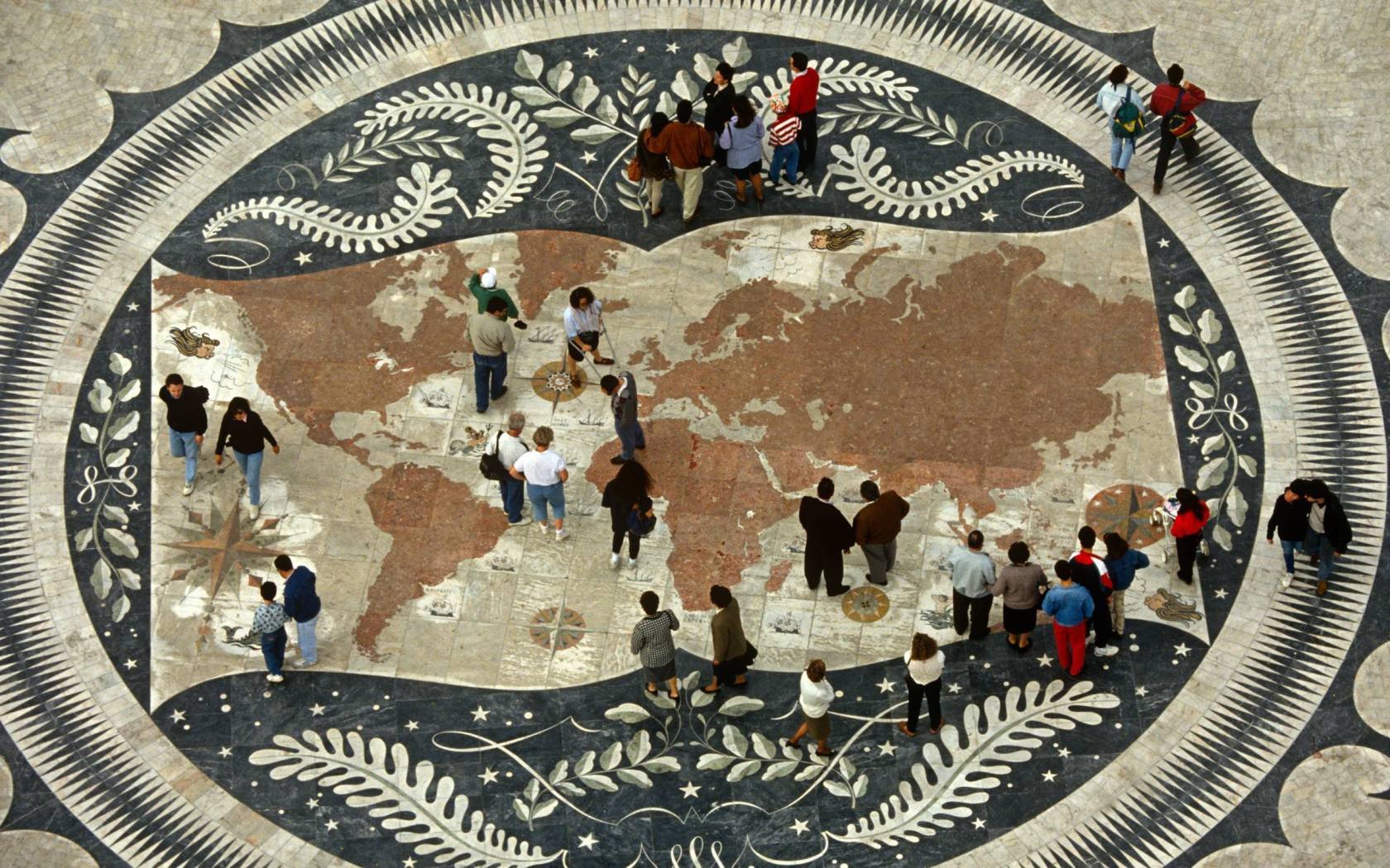Portuguese pedestrians walk over a world map on the pavement beneath the Monument of Discoveries, Lisbon. The world's landmass is represented here in a tiled mosaic that Portugal is famous for and citizens walk across this depiction of their planet like giants on a mini-sized map. Located in Bel��m, on the bank of the River Tagus where the monument celebrates an era of adventure, expansion and colonial ambition. Within a circular frame, the ornate map shows an almost ancient world minus its geopolitical borders.