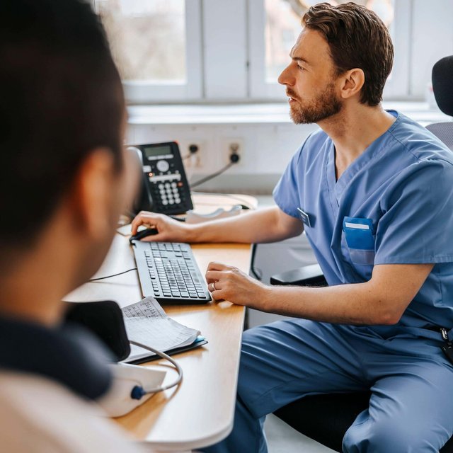 Focused male medical expert checking medical record of patient on computer while sitting on chair in examination room