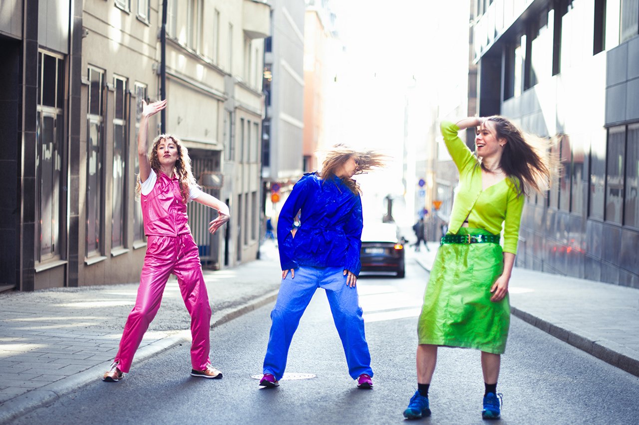 Tre kvinnor dansar mitt på en stadsgata i färgstarka kläder, omgivna av byggnader i dagsljus.