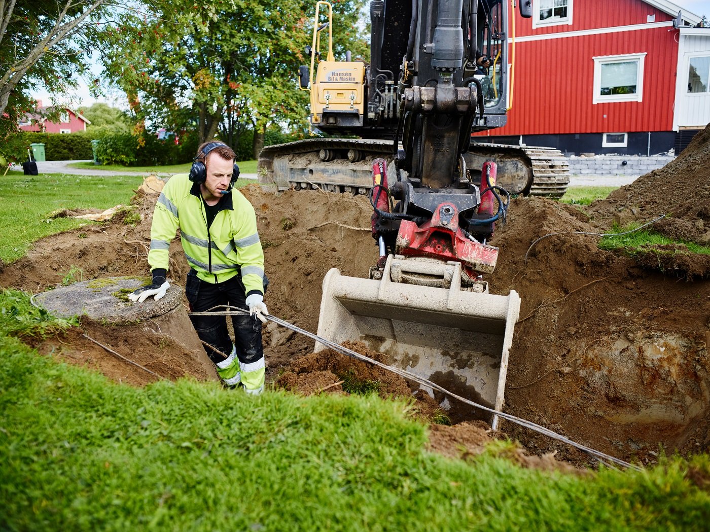 Arbete vid fastighet. Bilden visar en gr&auml;nvmaskin som gr&auml;ver och en man som hanterar en ledning.
