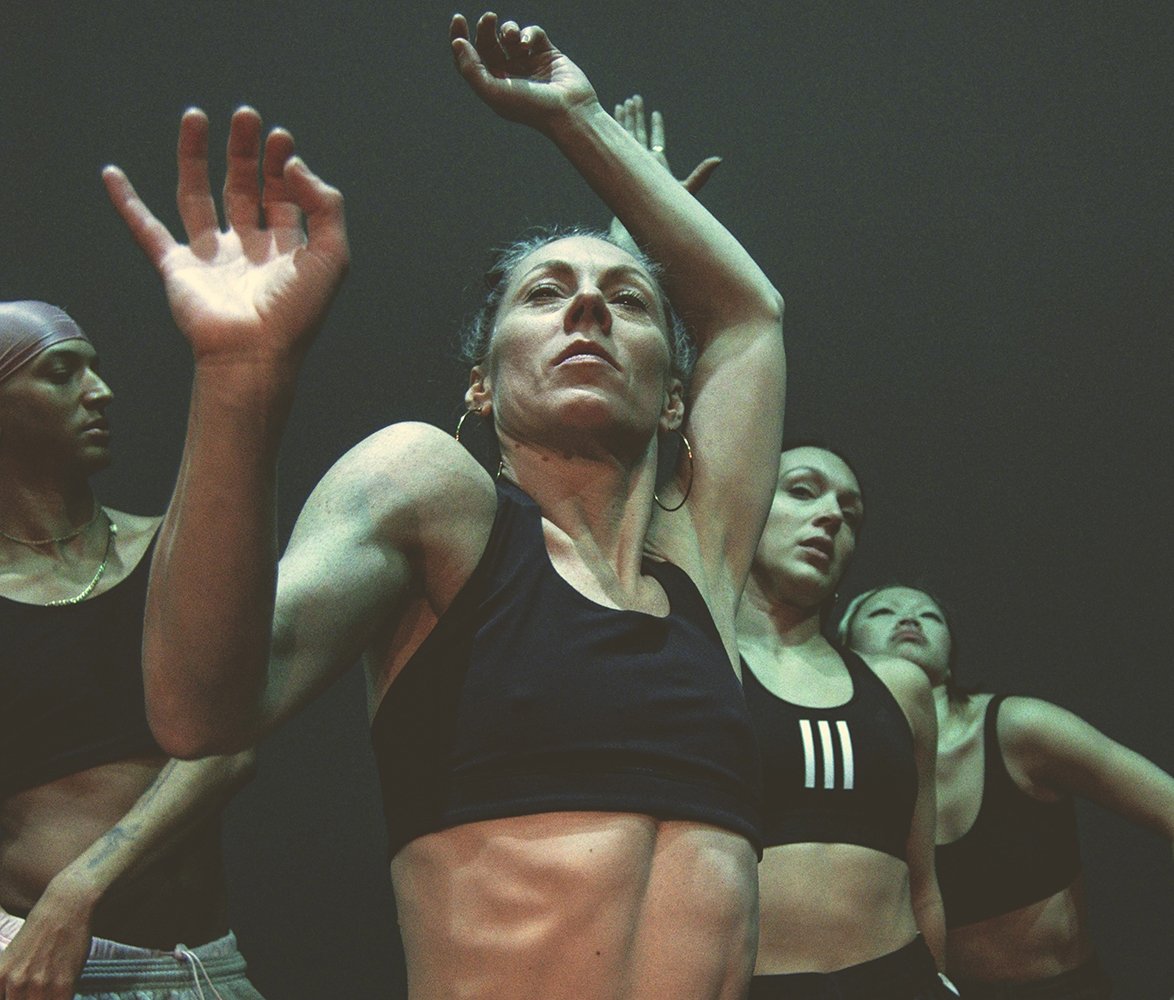 Fyra dansare står tätt tillsammans i en mörk scenmiljö. En person i förgrunden sträcker armarna uppåt medan de andra syns bakom i liknande poser. Bilden har ett intensivt, konstnärligt uttryck och fokus på kropp och rörelse.