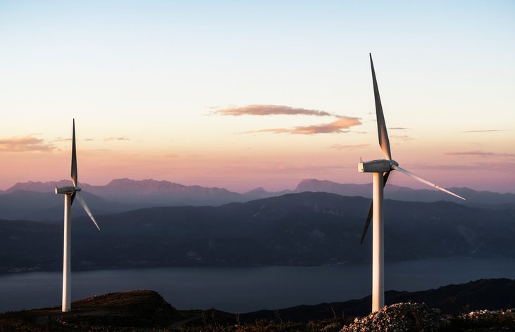 Two wind turbines stand out in front of a beautiful background of mountains during sunset.