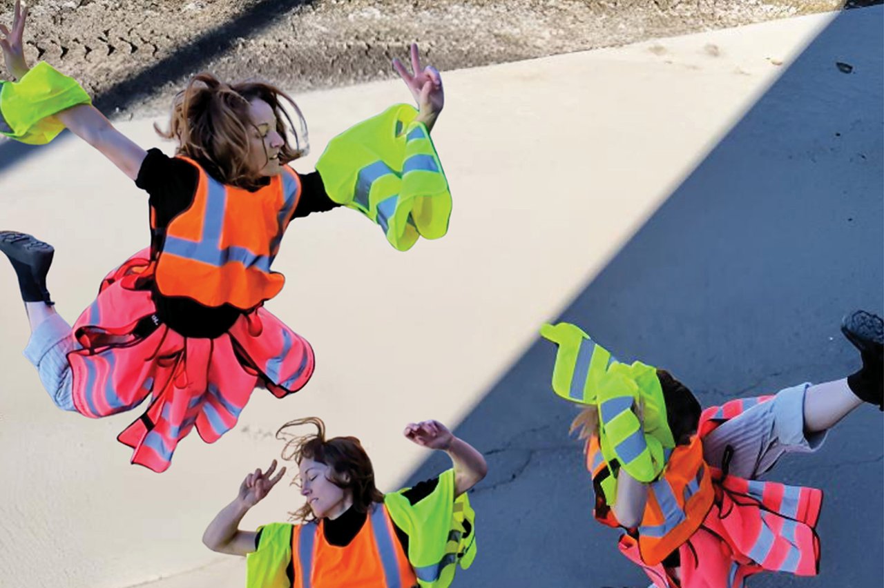 Dansare sedda ovanifrån, i rörelse på asfalt, klädda i orange och gula reflexvästar, fångade mellan ljus och skugga.
