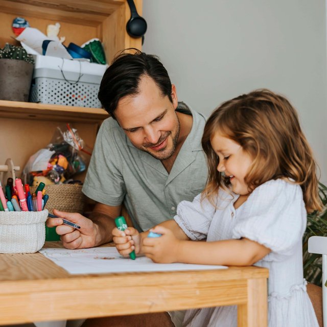 Father sitting next to daughter coloring with crayon while sitting at table