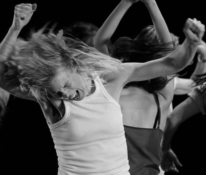 Kvinna med vildsint uttryck dansar med knutna nävar i luften, fler dansare syns i bakgrunden.