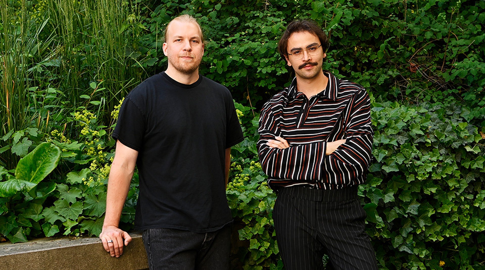 Afrang Nordlöf Malekian (höger) och Roger Smeby (vänster) står sida vid sida framför en mur med grönskande löv i bakgrunden, båda har fokuserade uttryck och blicken i kameran.