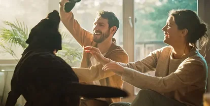 Om Mysafety försäkringar. Par leker med sin hund i vardagsrummet, vilket skapar en trygg och säker atmosfär hemma.