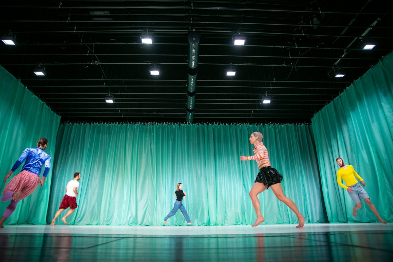 Flera dansare spridda över scenen, i rörelse mot olika riktningar framför en grön ridå.