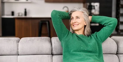 En eldre kvinne lener seg fornøyd tilbake i en grå sofa i huset sitt.