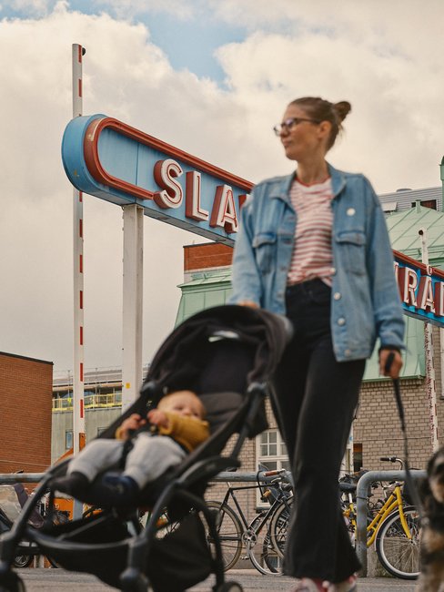 En kvinna g&aring;r med ett barn i en barnvagn. I bakgrunden syns skylten "Slakthusomr&aring;det".