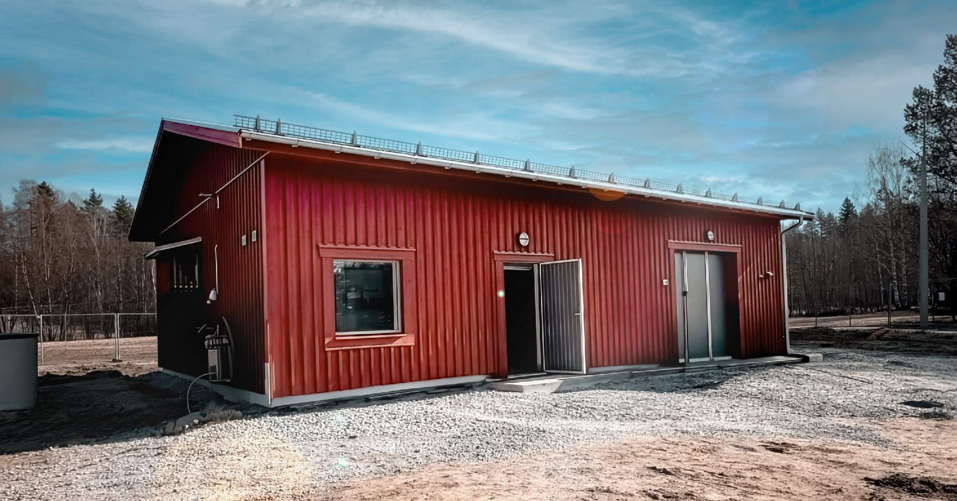 R&ouml;tt korrugerat industribyggnad med platta tak, f&ouml;nster och d&ouml;rrar, omgiven av grus och barrskog.