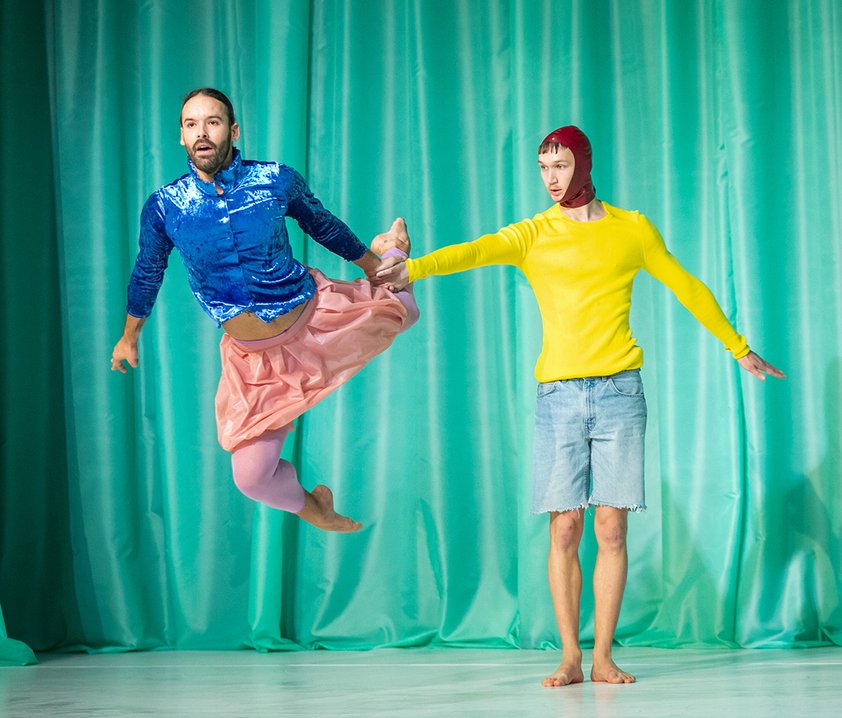 Två dansare i färgstarka kläder poserar mot en turkos bakgrund, där den ena svävar i ett hopp och den andra står stadigt.