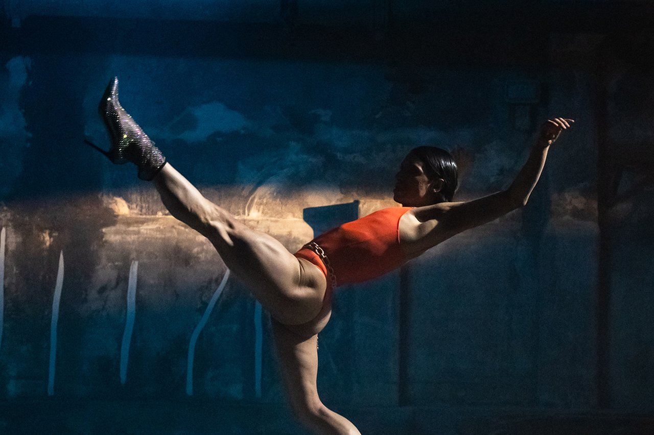 Dansaren Daniel Proietto står i röd dräkt och glittriga högklackade skor balanserar i en dramatisk danspose i ett dunkelt, industriellt rum med blått ljus.