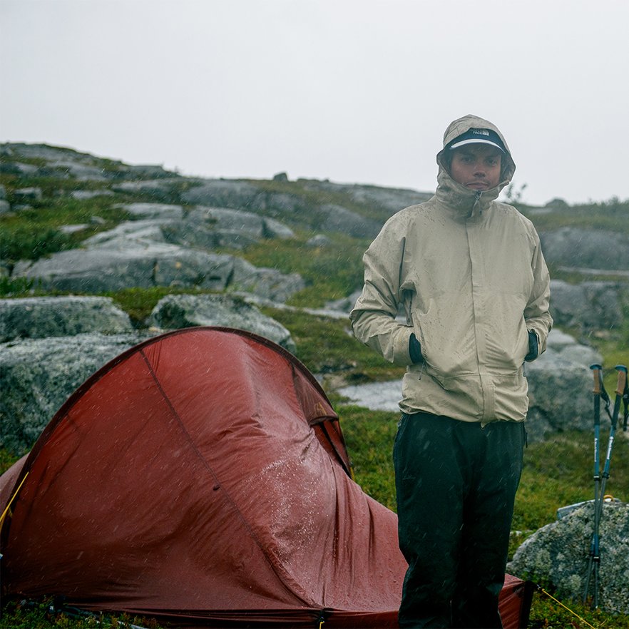 man st&aring;r med luvan uppdragen i regnet bredvid sitt r&ouml;da t&auml;lt