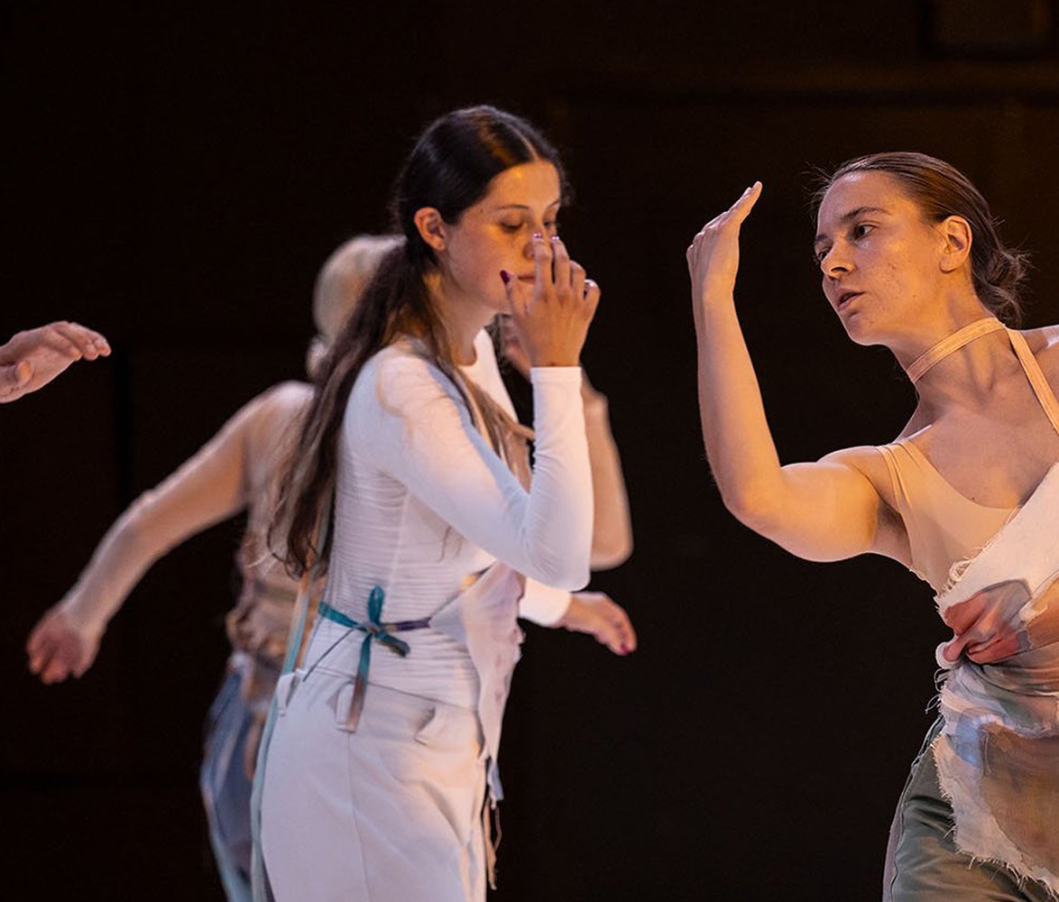 Tre personer dansar på en scen, klädda i ljusa, transparenta kostymer, med fokus på rörelse och samspel mellan kroppar.