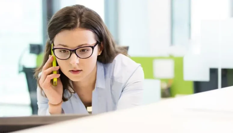 Nuori nainen toimistossa puhuu puhelimessa samalla kun katsoo tietokoneen näyttöä.