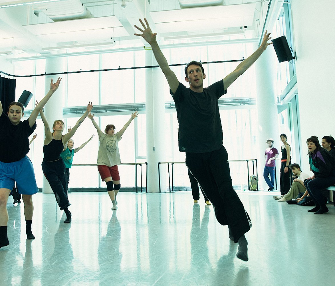 En grupp dansare tränar i en ljus dansstudio. I förgrunden rör sig en person framåt med armarna sträckta uppåt, medan flera andra dansare bakom speglar rörelsen. Längs väggen sitter några personer och tittar på repetitionen.