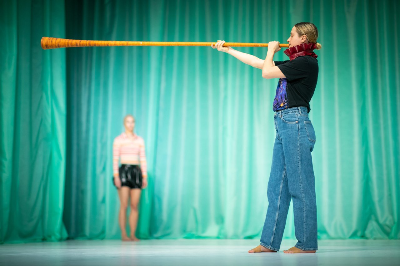 Dansare i förgrunden håller en lång stav horisontellt, med en annan dansare stående suddigt i bakgrunden.