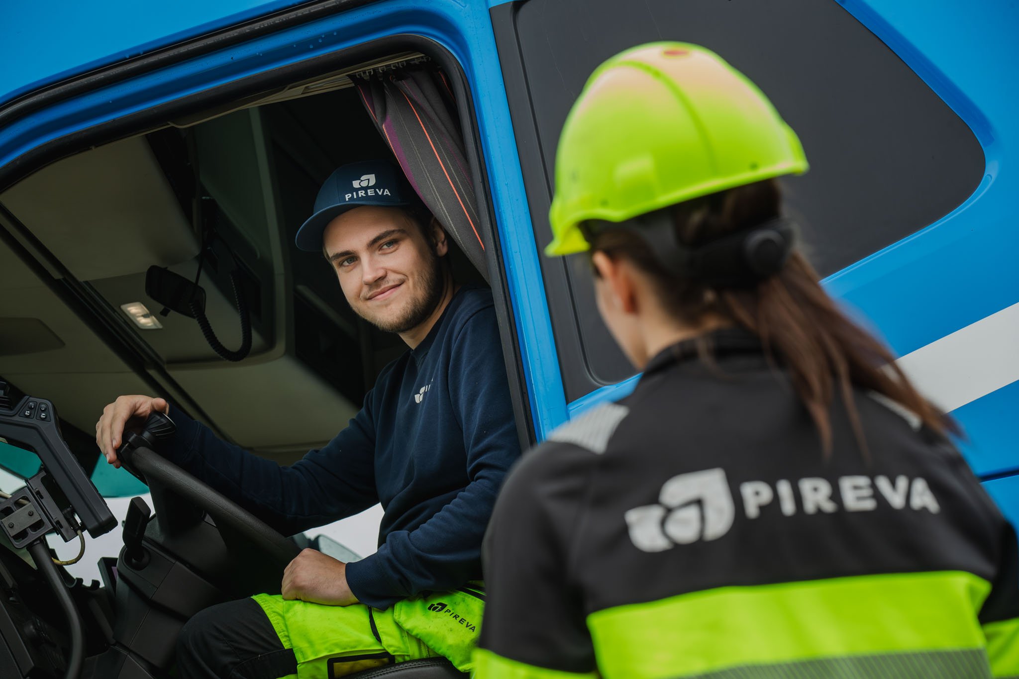 Pireva-arbetare i bl&aring; jacka sitter i f&ouml;rarhytten p&aring; en bl&aring; lastbil och talar med en kollega i gul s&auml;kerhetshjalm.