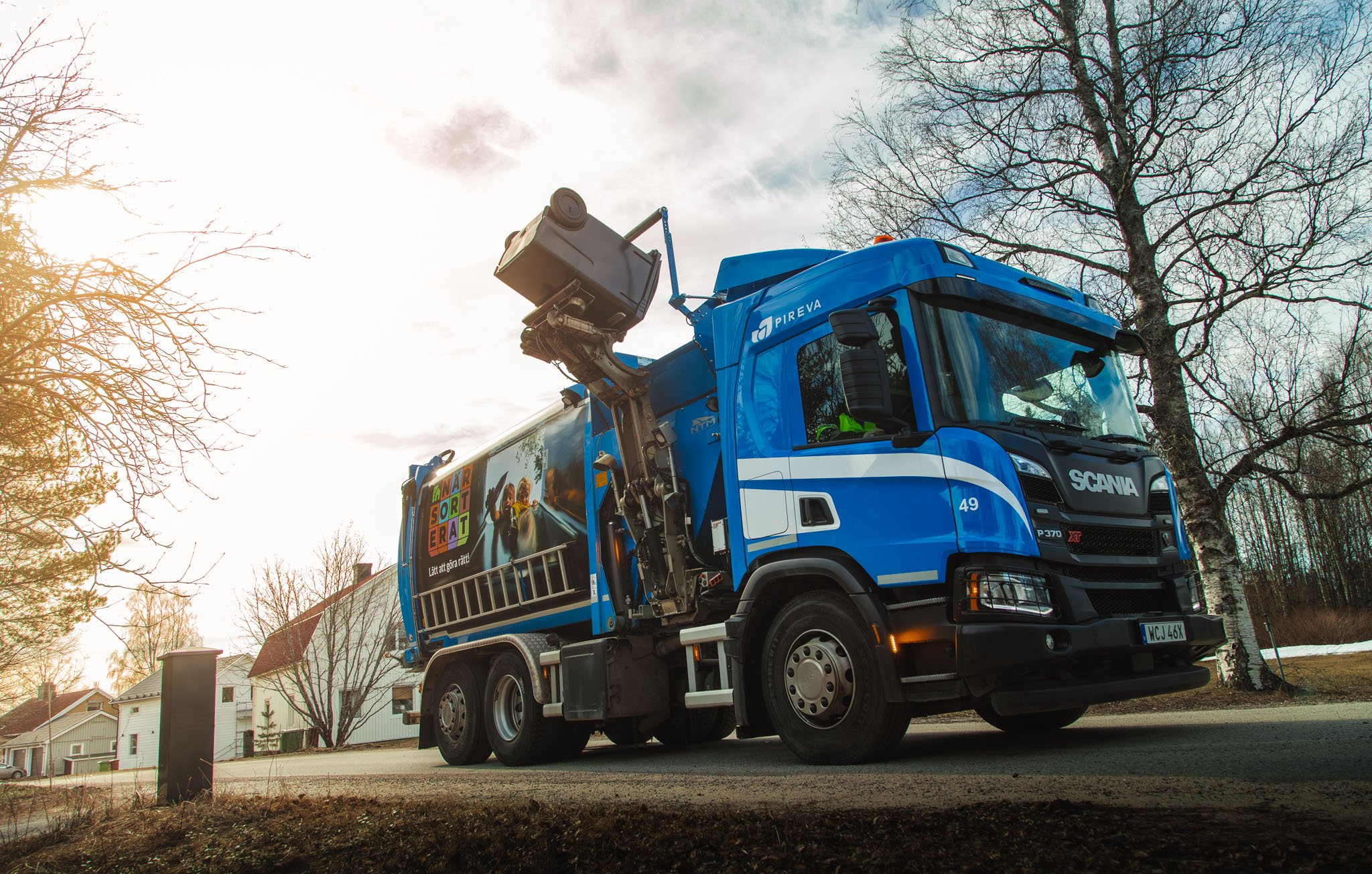 Bl&aring; Scania lastbil med sida-lastare, parkerad vid v&auml;g med tr&auml;d och hus.