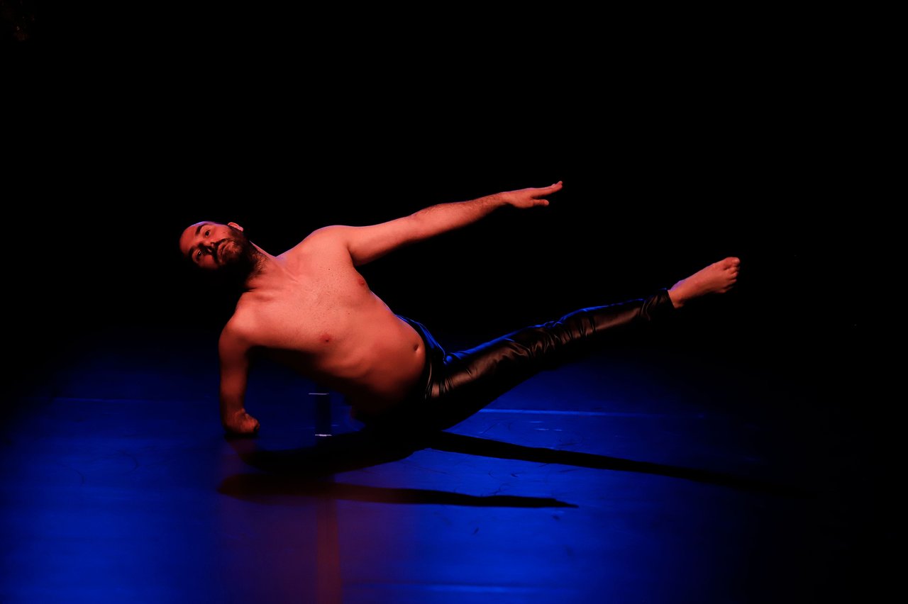 Dansare balanserar på ena armen med kroppen utsträckt, belyst i rött och blått mot svart bakgrund.