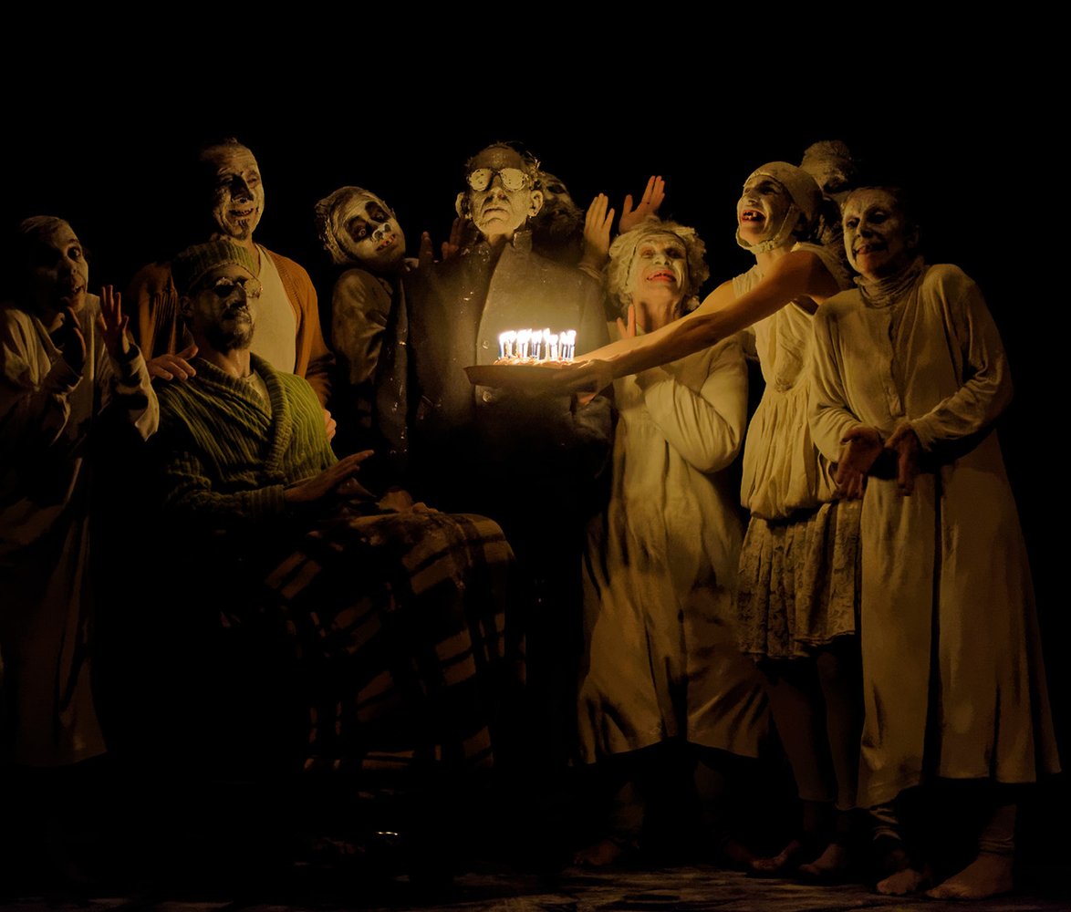 En grupp människor med vitsminkade ansikten, som räcker fram en födelsedagstårta mot en person.