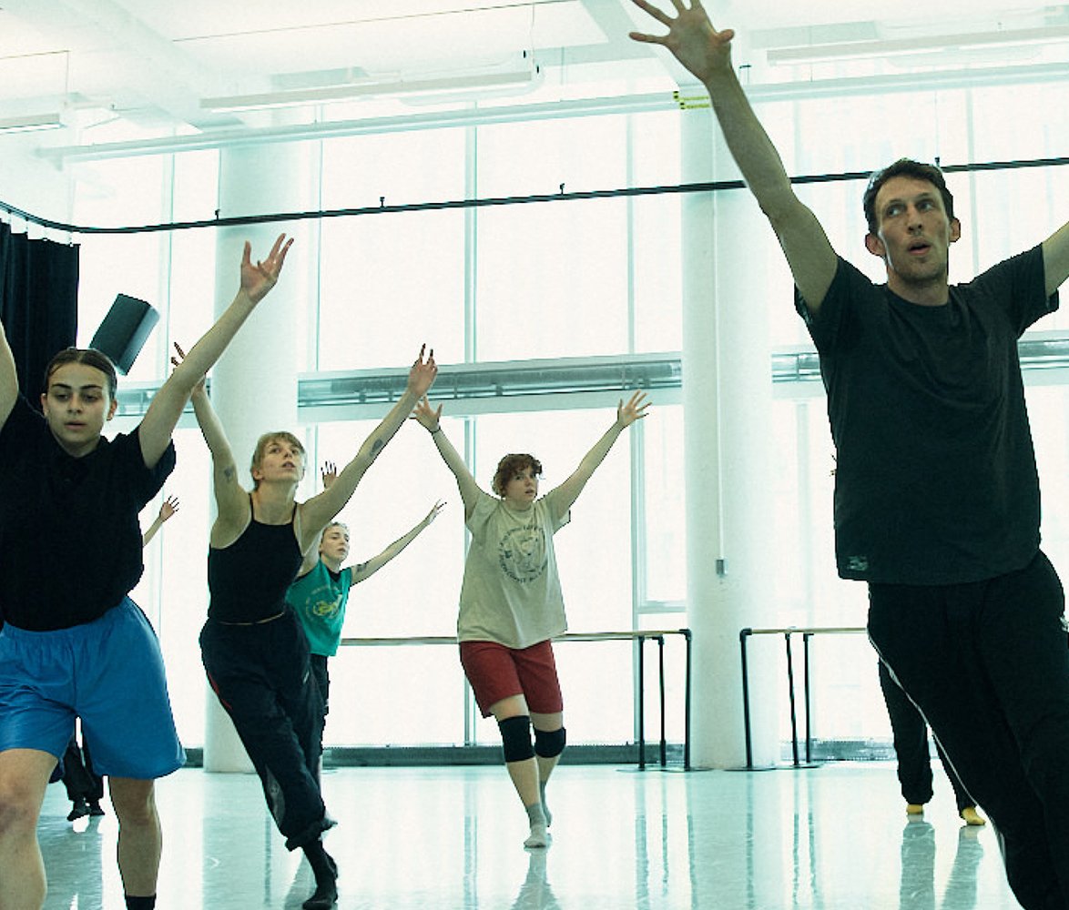 En grupp dansare tränar i en ljus dansstudio. I förgrunden rör sig en person framåt med armarna sträckta uppåt, medan flera andra dansare bakom speglar rörelsen. Längs väggen sitter några personer och tittar på repetitionen.