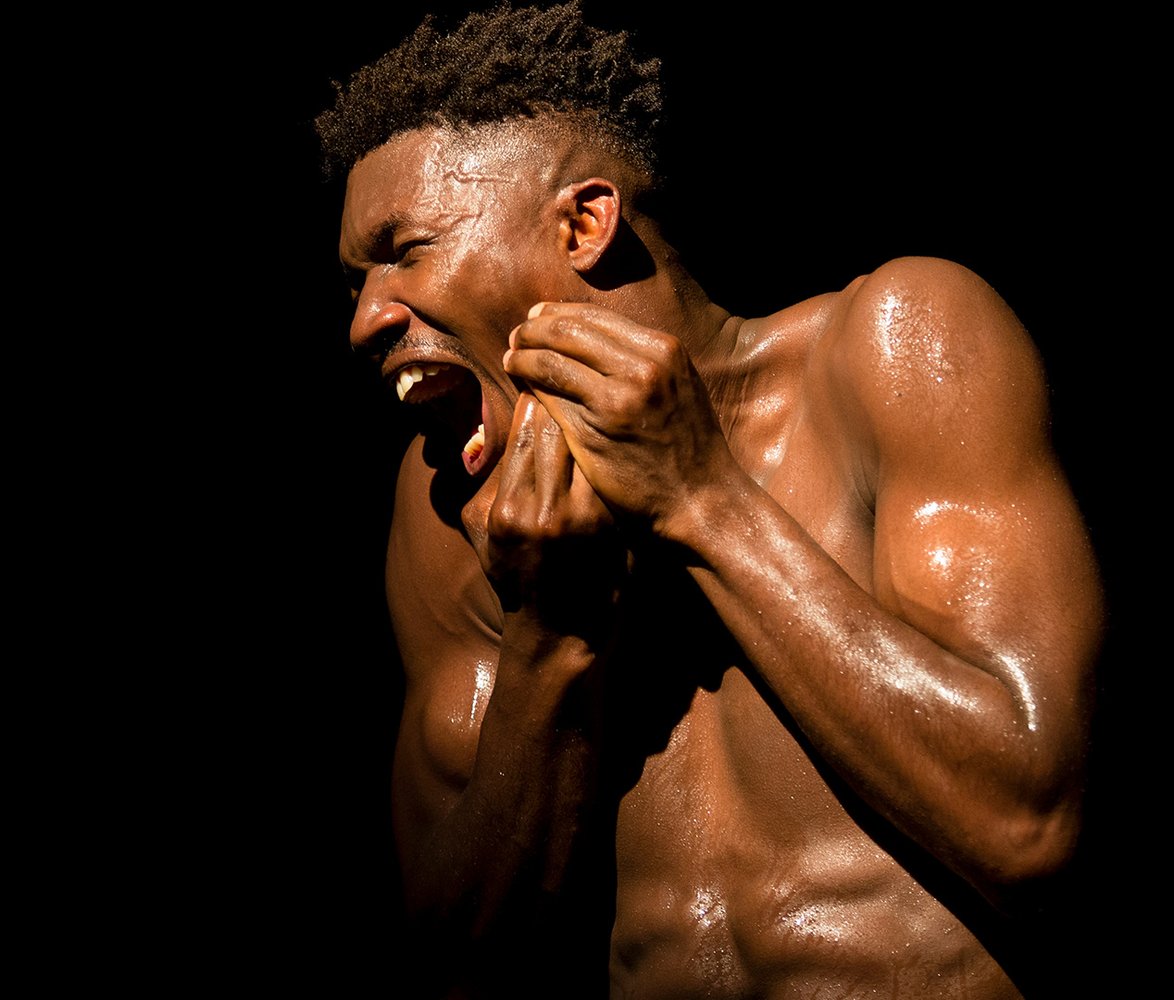 En svettig, barbröstad mörkhyad man med muskulös kropp uttrycker stark känsla genom ansikte och händer mot helt svart bakgrund.