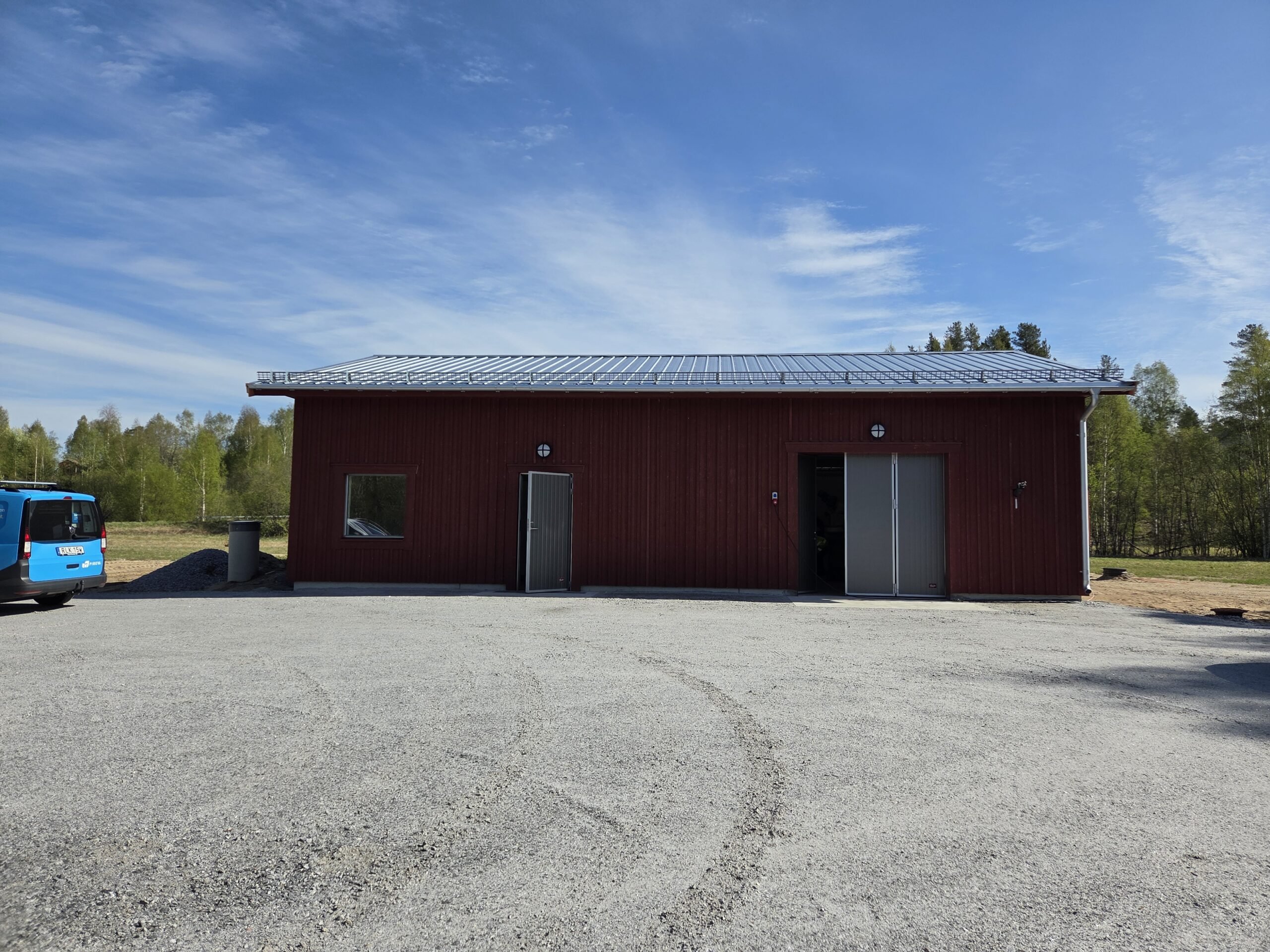 R&ouml;dbrunt industribyggnad med metalltak, tv&aring; d&ouml;rrar och f&ouml;nster. Grusplan framf&ouml;r. Skog och bl&aring; bil i bakgrunden.